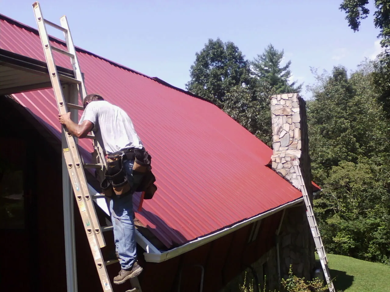 Roofer on a ladder beside a red metal roof and stone chimney — Soffit & Fascia Repair by Roofing Brothers in West Barnstable